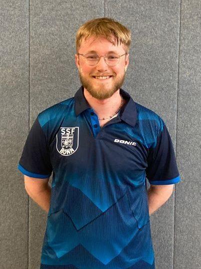 L&auml;chelnder junger Mann mit Brille tr&auml;gt ein blaues Tischtennis-Trikot vor einer grauen Wand.