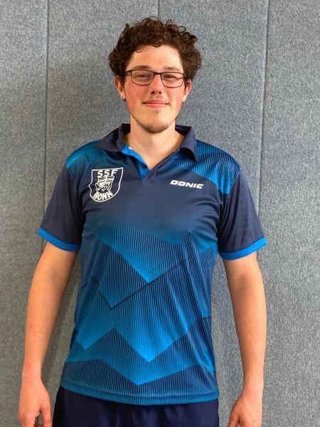 Junger Mann mit Brille tr&auml;gt ein blaues Sporttrikot mit geometrischem Muster und steht vor einer grauen Wand.