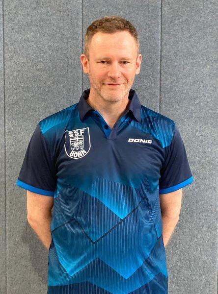 Mann in blauer Sportkleidung mit Vereinslogo steht vor grauer Wand.