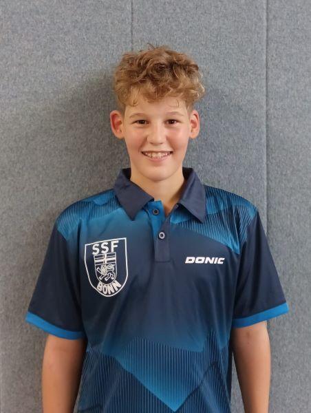 Portr&auml;t eines jungen Mannes mit lockigem Haar, der ein sportliches Polohemd des SSF Bonn tr&auml;gt. Hintergrund grau.