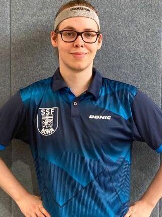 Junger Mann in einem blauen Tischtennistrikot mit Logo, steht vor einer grauen Wand und h&auml;lt seine H&auml;nde an der H&uuml;fte.