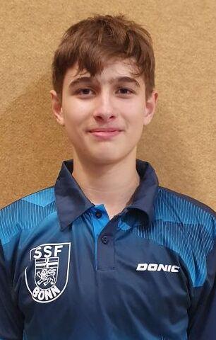 Portr&auml;t eines jungen Sportlers in blauem Tischtennisshirt mit Logo &bdquo;SSF Bonn&ldquo;, vor brauner Wand stehend.