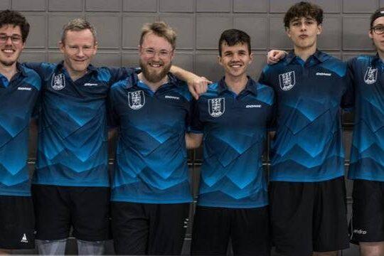 Sechs Tischtennisspieler in blauen Trikots stehen l&auml;chelnd nebeneinander vor einer grauen Wand.