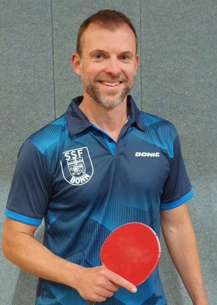 Lächelnder Mann in Sportkleidung hält einen roten Tischtennisschläger vor einer grauen Wand.
