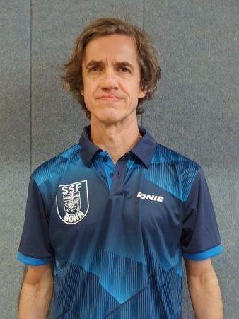 Mann mit dunklem, gestreiftem Sporthemd und Logo des SSF Bonn, vor grauer Wand stehend.