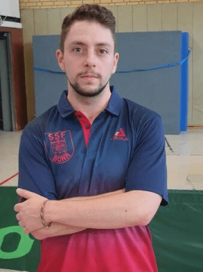 Junger Mann mit einem sportlichen Polo-Shirt steht in einer Sporthalle, Arme vor der Brust verschr&auml;nkt.