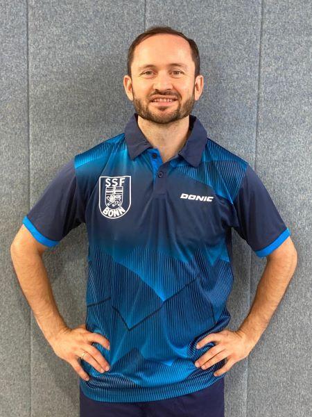 Mann in blauer Sportkleidung mit Vereinslogo steht l&auml;chelnd vor einer grauen Wand.