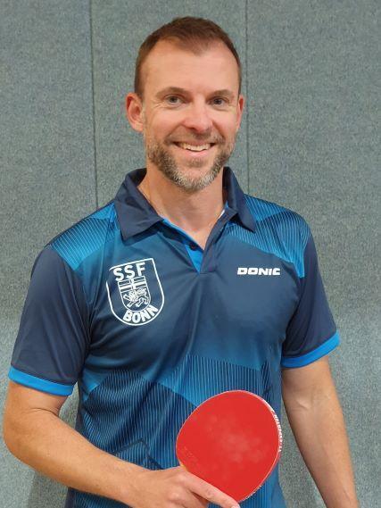 L&auml;chelnder Mann in Sportkleidung h&auml;lt einen roten Tischtennisschl&auml;ger vor einer grauen Wand.