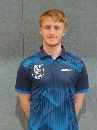 Junger Mann in blauer Sportkleidung mit Vereinslogo, steht l&auml;chelnd vor einer grauen Wand.