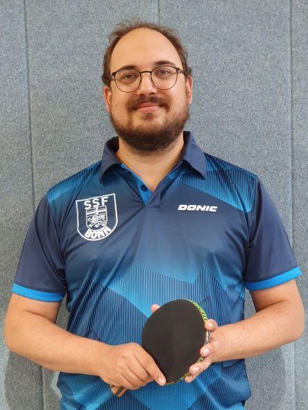 Mann mit Brille in Sportkleidung h&auml;lt einen Tischtennisschl&auml;ger vor einer grauen Wand.