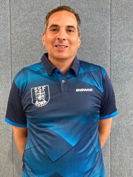 Mann in einem blauen Sporttrikot mit dem Logo "SSF Bonn" steht vor einer grauen Wand und l&auml;chelt.