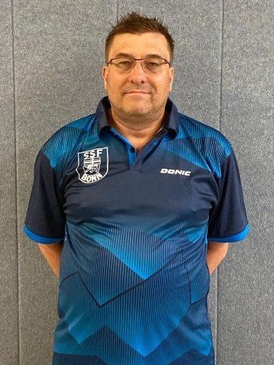 Mann in Sportshirt mit Vereinslogo steht vor grauem Hintergrund und l&auml;chelt.