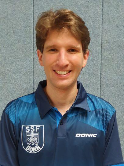 Portrait eines l&auml;chelnden Mannes in einem blauen Tischtennis-Trikot mit dem Aufdruck &bdquo;SSF BONN&ldquo; und &bdquo;DONIC&ldquo;.