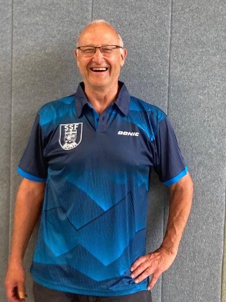 L&auml;chelnder Mann in einem blauen Sportshirt mit Logo vor grauer Wand.