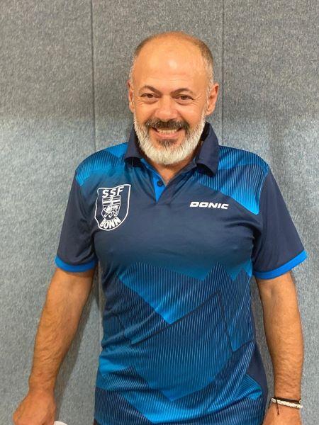 Mann mit Bart und l&auml;chelndem Gesicht tr&auml;gt ein blaues Tischtennis-Trikot vor einer grauen Wand.