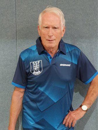 &Auml;lterer Mann in blauer Sportbekleidung mit Logo steht vor grauem Hintergrund.