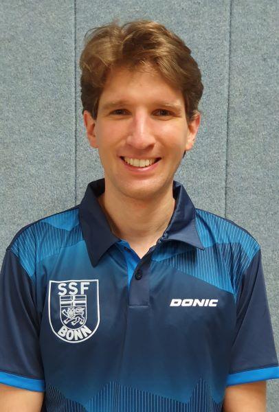 Portrait eines lächelnden Mannes in einem blauen Tischtennis-Trikot mit dem Aufdruck „SSF BONN“ und „DONIC“.