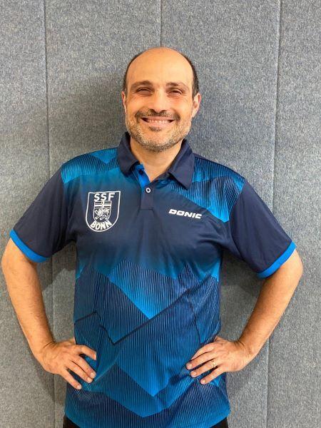 Mann mit kurzem Haar tr&auml;gt ein blaues Sporttrikot mit einem Wappen, l&auml;chelt und steht vor einer grauen Wand.