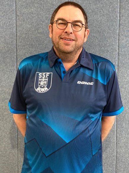 Mann in einem blauen Sporttrikot mit Logo, l&auml;chelnd vor einer grauen Wand.