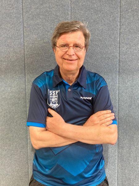 L&auml;chelnder Mann mit Brille in einem blauen Sportshirt mit Vereinslogo steht mit verschr&auml;nkten Armen vor einer grauen Wand.