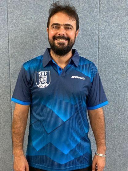 Mann mit Bart trägt ein blaues Sportshirt mit Logo und lächelt vor einer grauen Wand.