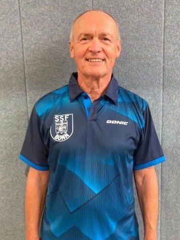 &Auml;lterer Mann in blauer Sportbekleidung mit Vereinslogo steht vor grauer Wand.