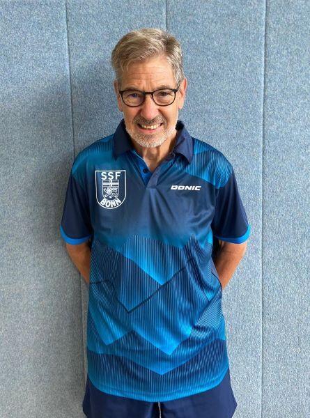 Mann mit Brille in blauer Sportkleidung steht vor einer grauen Wand.