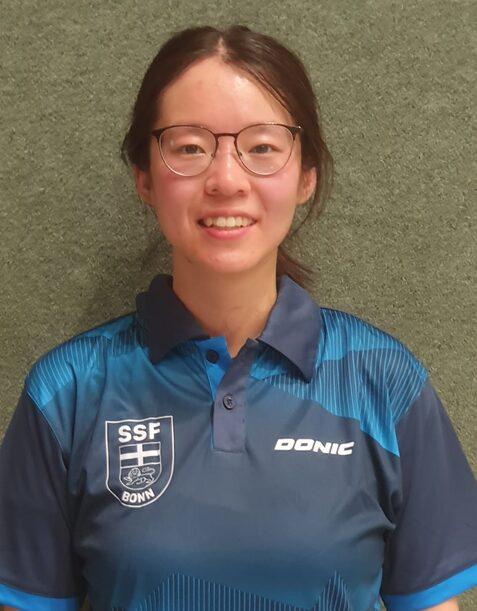 Porträt einer lächelnden Person mit Brille, getragenem Sporttrikot der SSF Bonn in blau, vor einer grünen Wand.