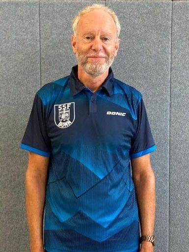Mann in blauer Sportkleidung mit Logo steht vor einer grauen Wand.