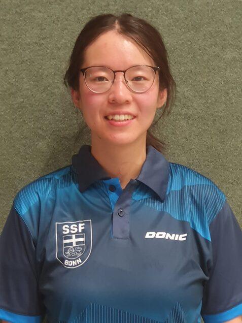 Portr&auml;t einer l&auml;chelnden Person mit Brille, getragenem Sporttrikot der SSF Bonn in blau, vor einer gr&uuml;nen Wand.