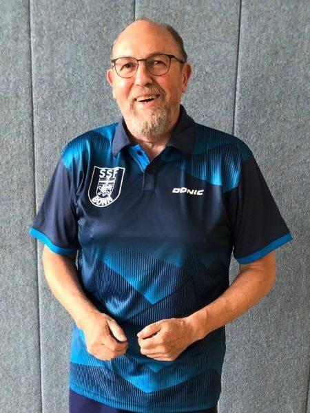 L&auml;chelnder Mann in blauer Sportkleidung mit Vereinslogo steht vor grauem Hintergrund.