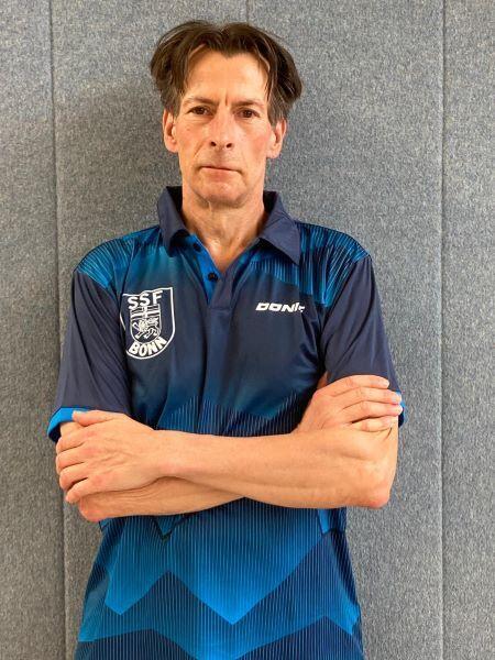 Mann in blauer Sportkleidung mit Logo steht mit verschr&auml;nkten Armen vor grauer Wand.