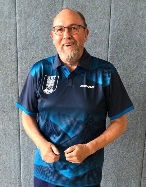 Lächelnder Mann in blauer Sportkleidung mit Vereinslogo steht vor grauem Hintergrund.