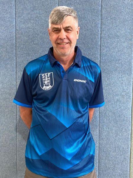 Mann in blauem Sportshirt mit Vereinslogo steht vor grauer Wand.