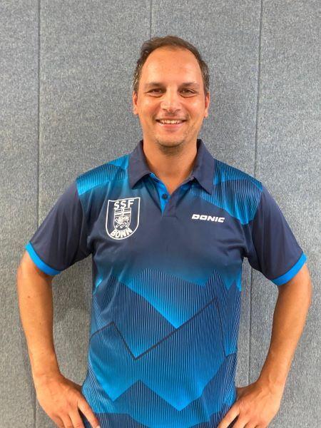 L&auml;chelnder Mann in sportlichem, blauem Shirt mit Vereinslogo, stehend vor grauer Wand.