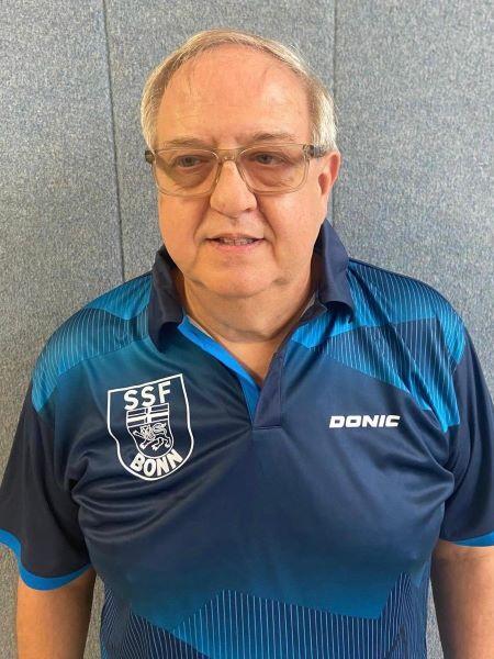 Portr&auml;t eines Mannes in einem blauen Sporthemd mit dem Logo &bdquo;SSF BONN&ldquo; und Brille, vor einer grauen Wand.