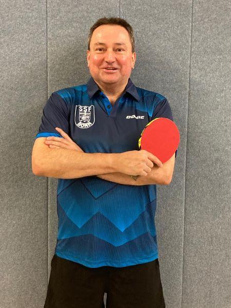 Mann in blauem Tischtennisshirt h&auml;lt einen Tischtennisschl&auml;ger und l&auml;chelt vor einer grauen Wand.