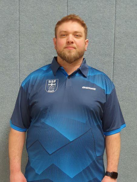 Ein Mann mit Bart tr&auml;gt ein blaues Sporthemd mit Logos auf der Brust und posiert vor einer grauen Wand.