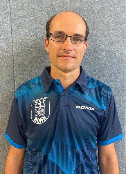 Mann in Sportkleidung mit SSF Bonn-Logo steht vor einer grauen Wand.