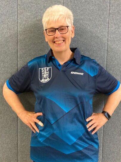 Frau mit kurzen, grauen Haaren und Brille tr&auml;gt ein blaues Tischtennis-Trikot und l&auml;chelt vor einer grauen Wand.