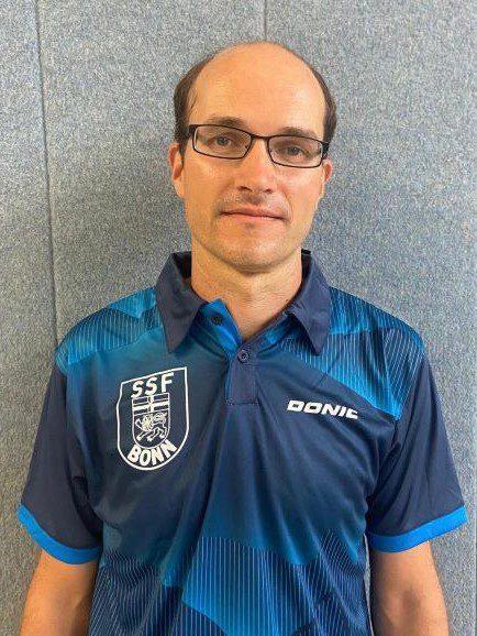 Mann in Sportkleidung mit SSF Bonn-Logo steht vor einer grauen Wand.