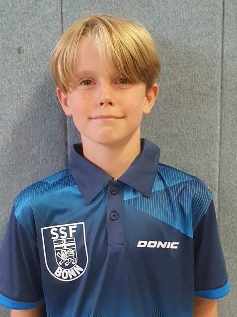 Junger Junge mit blonden Haaren tr&auml;gt ein blaues Sportshirt mit dem Logo &bdquo;SSF Bonn&ldquo; und steht vor einer grauen Wand.
