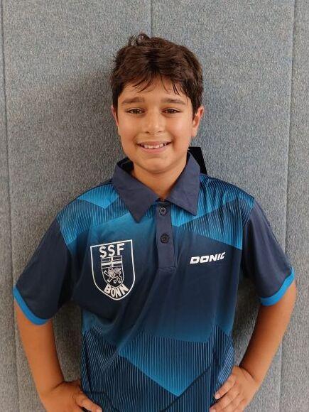 L&auml;chelnder Junge in blauer Tischtenniskleidung mit Logo, steht vor grauem Hintergrund.