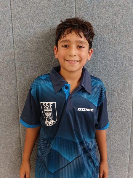 Junge mit dunklen Haaren tr&auml;gt ein blaues Tischtennistrikot vor einer grauen Wand. L&auml;chelt freundlich in die Kamera.