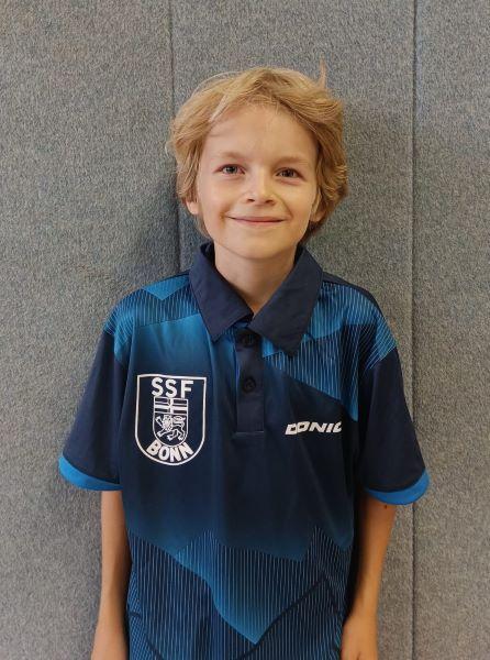 L&auml;chelnder Junge mit blonden Locken in einem blauen Tischtennis-Trikot vor einer grauen Wand.