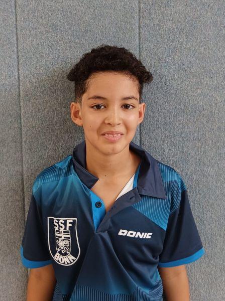 Ein junger Sportler in blauem Tischtennis-Trikot l&auml;chelt vor einer grauen Wand.