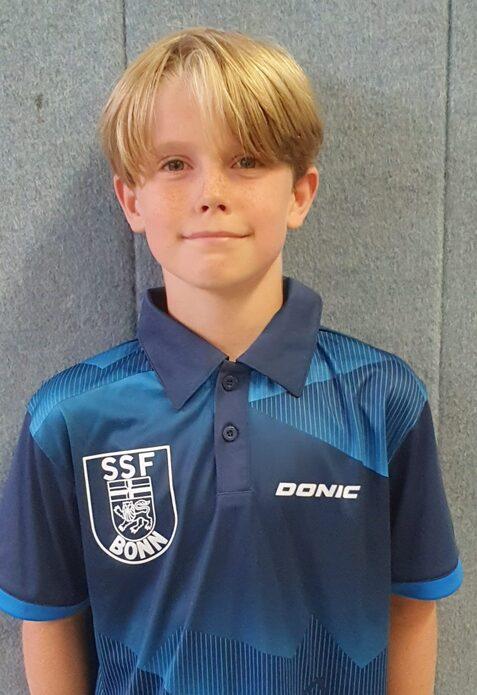 Junger Junge mit blonden Haaren trägt ein blaues Sportshirt mit dem Logo „SSF Bonn“ und steht vor einer grauen Wand.