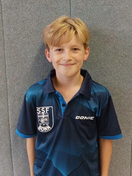 Junger Junge mit blondem Haar l&auml;chelt in einem blauen Tischtennis-Trikot vor einer grauen Wand.