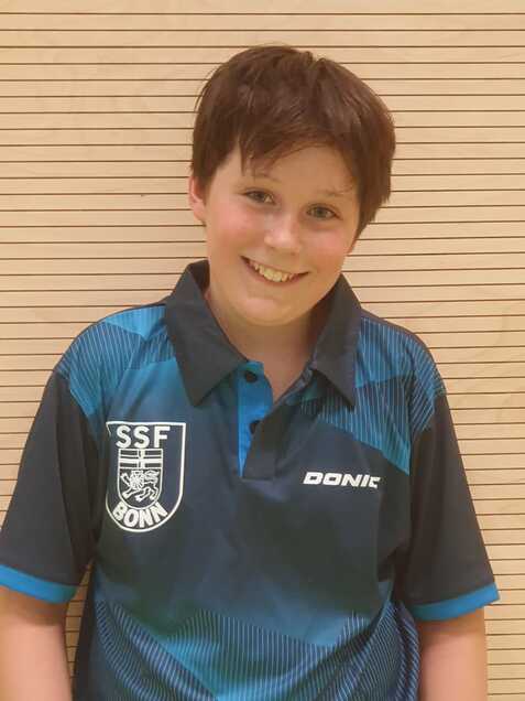 Junge in T-Shirt mit SSF Bonn-Logo l&auml;chelt vor einer hellen Wand aus Holzlatten.