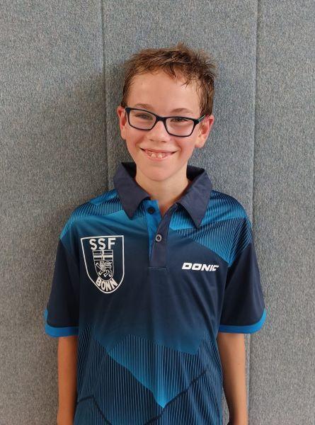 L&auml;chelnder Junge mit Brille tr&auml;gt ein blaues Trikot mit SSF-Bonn-Logo vor einer grauen Wand.